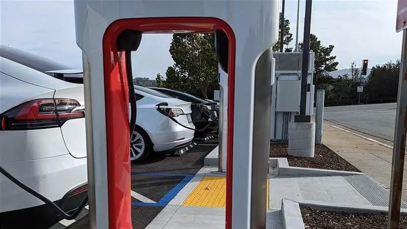 White Tesla Supercharger charging post with a red cable cradle at a station in San Mateo, California, with Tesla cars plugged into adjacent stalls