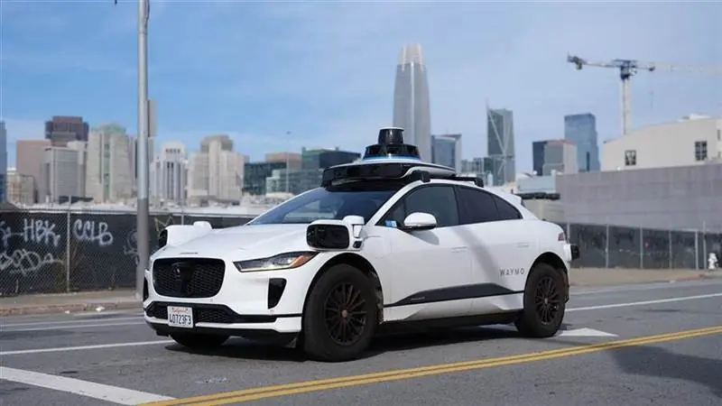 White Waymo Jaguar I-Pace robotaxi with roof-mounted sensor pod driving on a San Francisco street, city skyline in the background